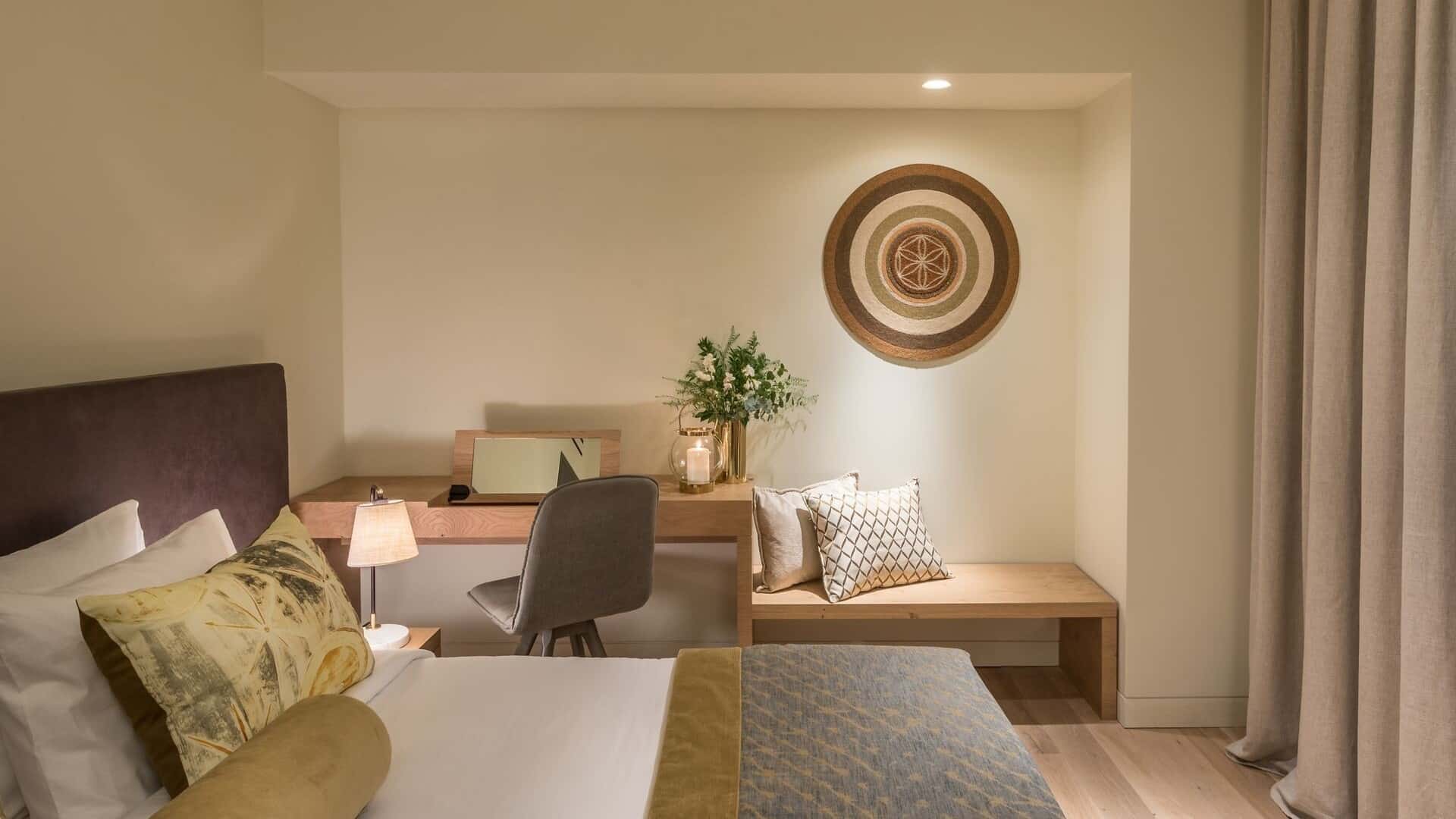 The interiors of our Classic Room, dressed in beige and earth tones, showing the bed and writing desk with bench beside it.
