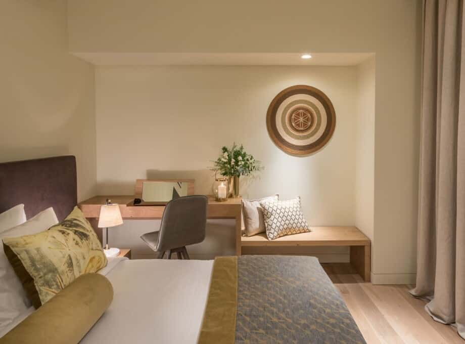 The interiors of our Classic Room, dressed in beige and earth tones, showing the bed and writing desk with bench beside it.