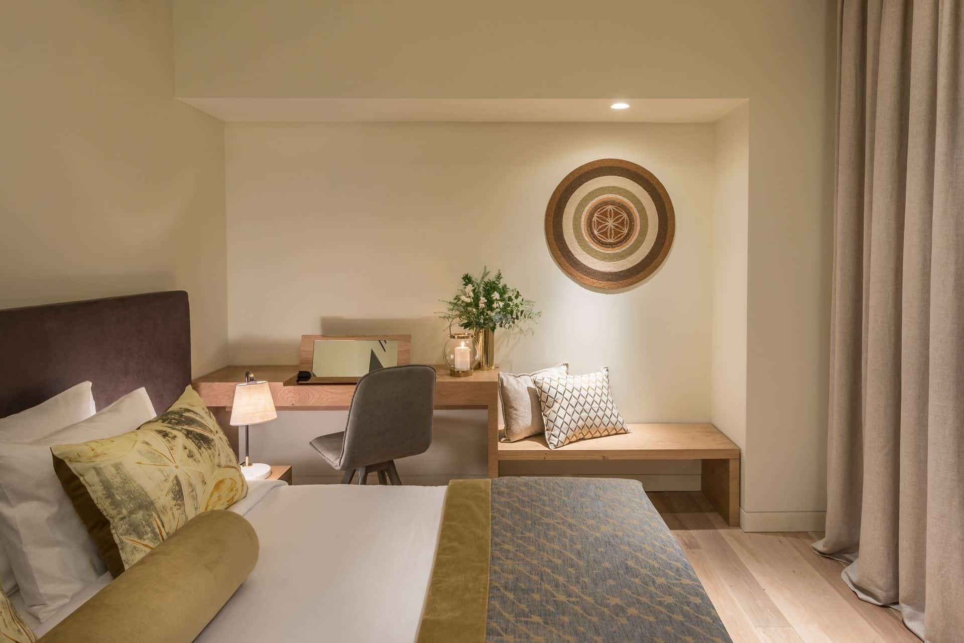 The interiors of our Classic Room, dressed in beige and earth tones, showing the bed and writing desk with bench beside it.
