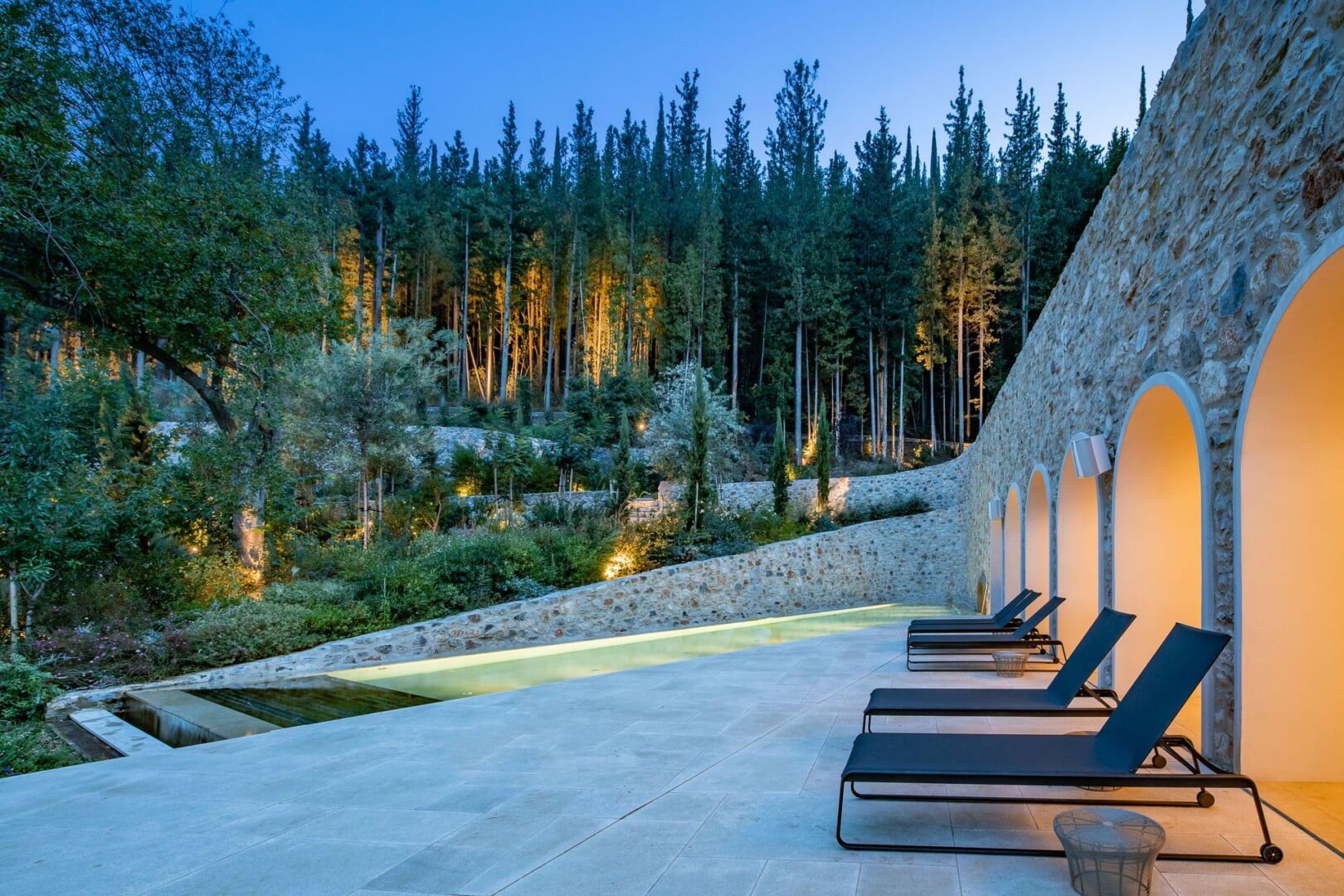 The pool terrace at dusk, the water glowing green between arched recesses in the stone wall and a pine forest lit softly up the slope behind.