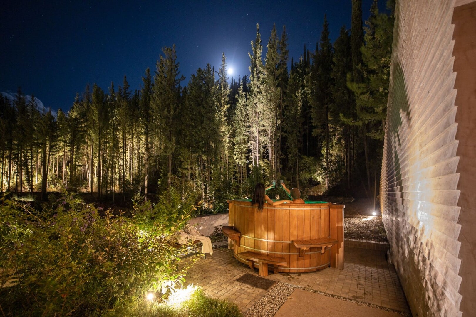 Night capture of the outdoor hot tub at Euphoria Retreat with people relaxing, under the moonlight, surrounded by a forest.