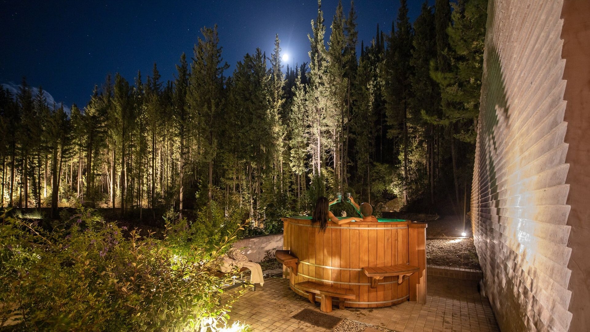 Night capture of the outdoor hot tub at Euphoria Retreat with people relaxing, under the moonlight, surrounded by a forest.