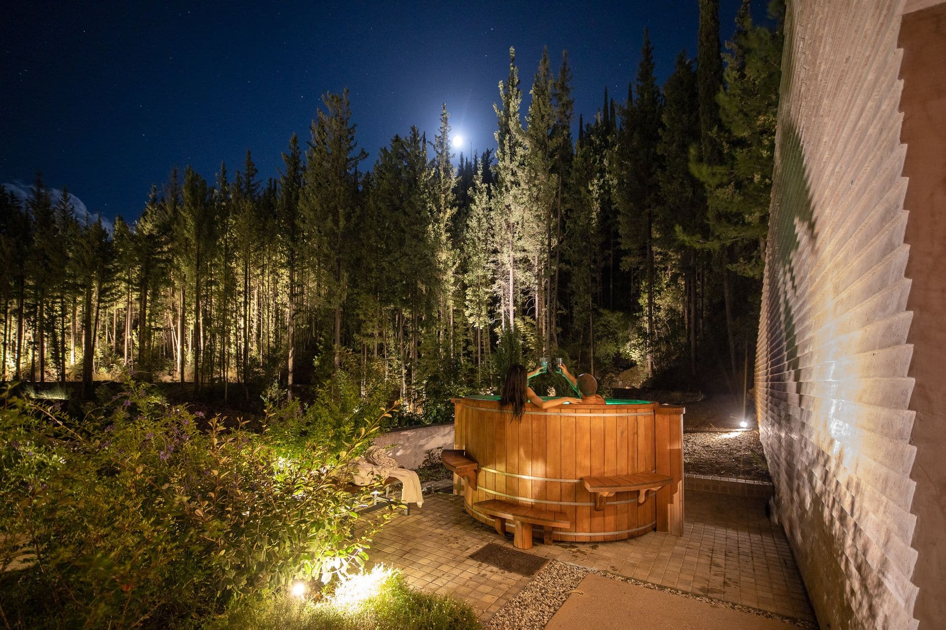 Night capture of the outdoor hot tub at Euphoria Retreat with people relaxing, under the moonlight, surrounded by a forest.