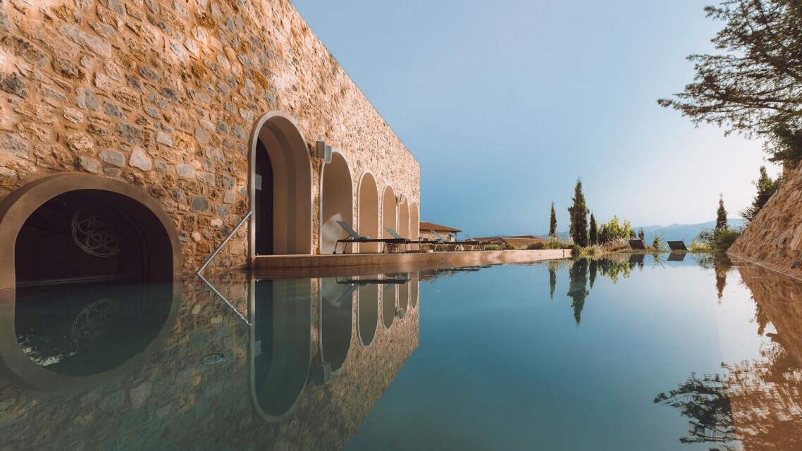 An eye-level outdoor shot shows the stone building of the Euphoria Retreat with arched windows and doors reflected in a still, clear infinity pool under a bright blue sky.