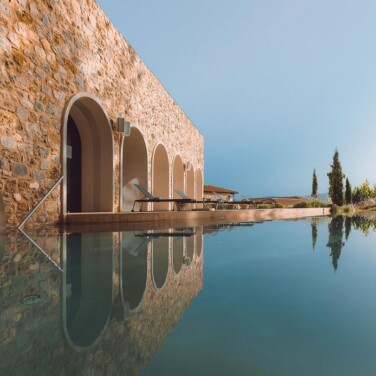 An eye-level outdoor shot shows the stone building of the Euphoria Retreat with arched windows and doors reflected in a still, clear infinity pool under a bright blue sky.